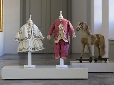 Kleidchen mit Paletot-Jacke für einen Jungen oder ein Mädchen, weißer Baumwoll-Piqué mit schwarzer Stickerei, Frankreich um 1860 / Höfischer Anzug für einen etwa 4-jährigen Jungen rosa Seide aus Lucca, 1790er Jahre / Reitpferd zum Ziehen, 19. Jh. (um 1850)