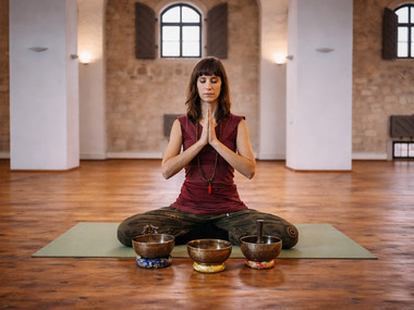 Ein Frau sitzt in Meditationshaltung auf einer Yoga-Matte, vor ihr stehen KLangschalen