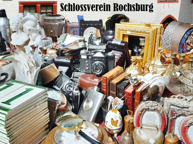 Plakat zum Flohmarkt mit Datum- und Zeitangaben. Im Hintergrund Fassade von Schloss Rochsburg. Im Vordergrund ein Tisch mit verschiedenen Flohmarktprodukten, z.B. Kameras, Porzellan, Kerzenständer