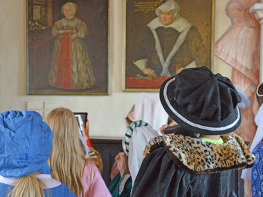 Das Bild zeigt Kinder in nachgeschneiderten historischen Kostümen, wie sie im Festsaal von Schloss Rochsburg die Ahnengalerie der Familie von Schönburg betrachten. 