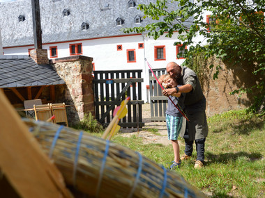 Ritter mit Pfeil und Bogen zeigt einem Jungen das Bogenschießen, im Hintergrund Burghof von Schloss Rochsburg